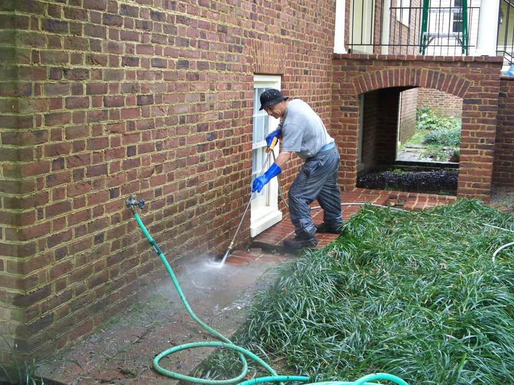 Person pressure washing a brick patio near a house; green hose is connected to a faucet on the brick wall.