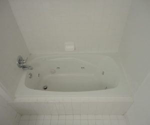 White bathtub with jets, surrounded by white tile walls, a soap holder, and a silver faucet on the left side.