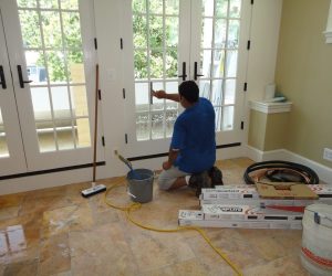 A person kneels and cleans glass doors in a sunlit room with cleaning supplies and boxes on the tiled floor.