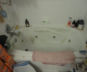 Dirty white bathtub with various bottles on the rim, a pink towel draped over the edge, and cleaning supplies nearby.