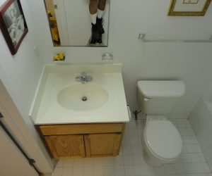 Bathroom with white sink, wooden cabinet, toilet, and mirror reflecting a person’s legs and shoes.