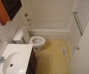 Small bathroom with a bathtub, toilet, sink, floral wallpaper, yellow flooring, and a towel bar on the wall.