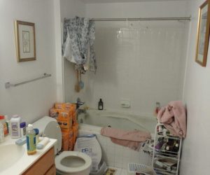 Cluttered bathroom with toiletries, boxes, and laundry scattered on floor and surfaces, towel hanging on shower curtain rod.