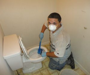 Person wearing gloves and a mask scrubbing a toilet in a small bathroom with stained floor tiles.
