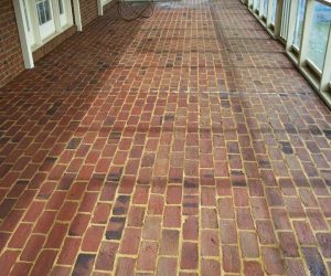 Red brick floor on a covered patio with glass windows on the right and a brick wall with doors on the left.
