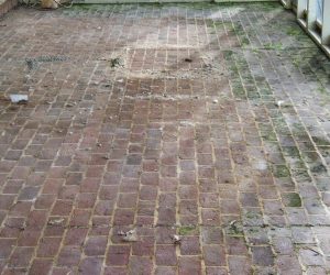 Red brick patio with some dirt patches and worn bricks, surrounded by a partial fence or railing.