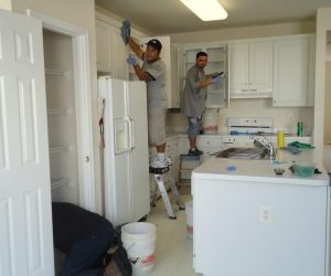 Three people clean a kitchen; two stand on stools to reach cabinets while one works near the pantry.