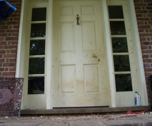 A dirty white front door with sidelights, set in a brick wall; debris is scattered on the brick porch.