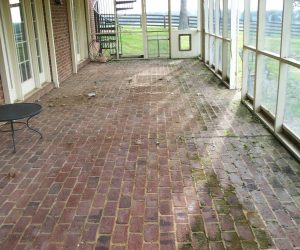 Screened porch with brick floor, some moss and dirt patches, round table, and view of a fenced yard in the background.