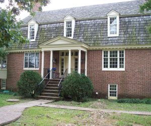 Two-story brick house with wood shingle roof, central steps, columns, and three dormer windows on upper floor.