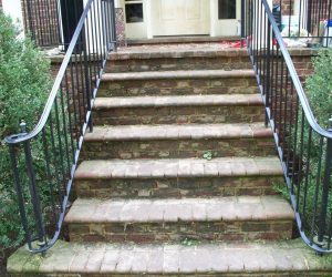 Worn brick steps with black metal railings, leading up to a double-door entrance, flanked by green bushes.