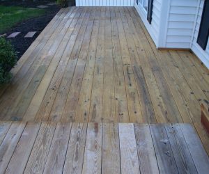 A wooden deck with weathered and discolored planks adjacent to a white house and a garden area.