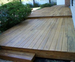 Wooden deck with two levels and a small step, adjacent to a house, surrounded by bushes and a white fence.