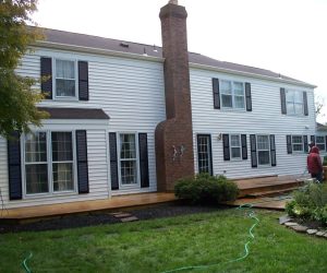 Two-story white house with black shutters, brick chimney, wooden deck, bushes, and a person standing outside.