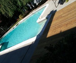 A backyard swimming pool with clear water bordered by a wooden deck and concrete patio, partly in shadow.