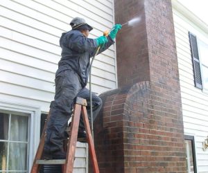 Person on ladder pressure washing a brick chimney attached to the exterior wall of a house.