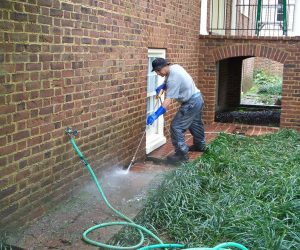 Person pressure washing a brick patio near a house; green hose is connected to a faucet on the brick wall.