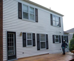 Person pressure washing a wooden deck outside a white house with black shutters and multiple windows.