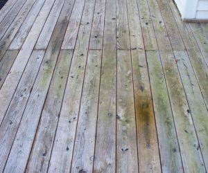 Weathered wooden deck boards with visible discoloration and knots next to the siding of a white house.
