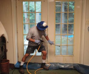Person in gloves and hat uses a pressure washer to clean glass doors indoors, with plastic sheets on the floor.