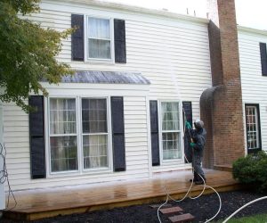 Person power washing dirt and mildew from the exterior siding of a two-story house with black shutters.
