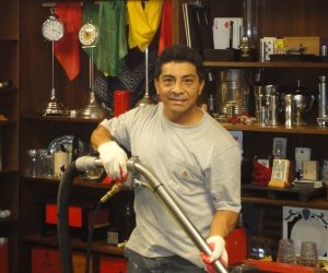 Man in gray shirt and gloves vacuuming in a room with shelves of glassware, clocks, and assorted objects.