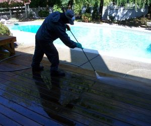 Person pressure washing a wooden deck beside a swimming pool on a sunny day.
