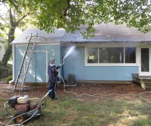 Person using a pressure washer to clean the exterior wall of a blue house; ladder and equipment visible nearby.