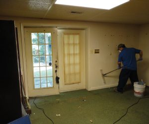 Person cleaning a wall in a mostly empty room with green carpet, near a door with a window and sheer curtain.
