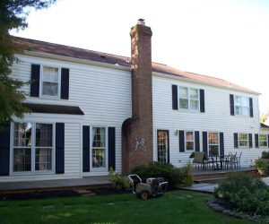 Two-story white house with black shutters, brick chimney, patio furniture, and a wheelbarrow on the lawn.