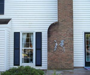 A brick chimney on a white house has damaged bricks and missing mortar forming irregular shapes on its surface.