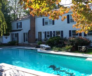 A backyard swimming pool in front of a two-story white house with patio furniture and surrounding greenery.