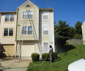 Person pressure washing dirt off the exterior of a beige townhouse on a sunny day, with ladders leaning on the house.