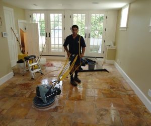 Person cleaning tile floor with a large floor buffer in a bright, unfurnished room with cleaning supplies visible.