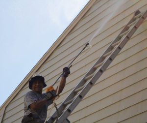 Person using a pressure washer to clean the siding of a house while standing on a ladder.