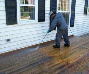 Person in gray clothing power washes a wooden deck beside a white house with black shutters and windows.