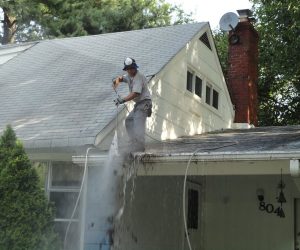A man power washing debris off a house roof, with water and dirt falling to the ground below.