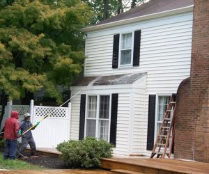 Two people use a pressure washer to clean the exterior siding of a white house near a brick chimney.