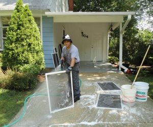 Person washing window screens with a hose and soap on a driveway in front of a house.