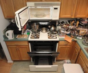 Cluttered kitchen with an open oven, pots on the stove, open microwave, and countertops filled with various items.